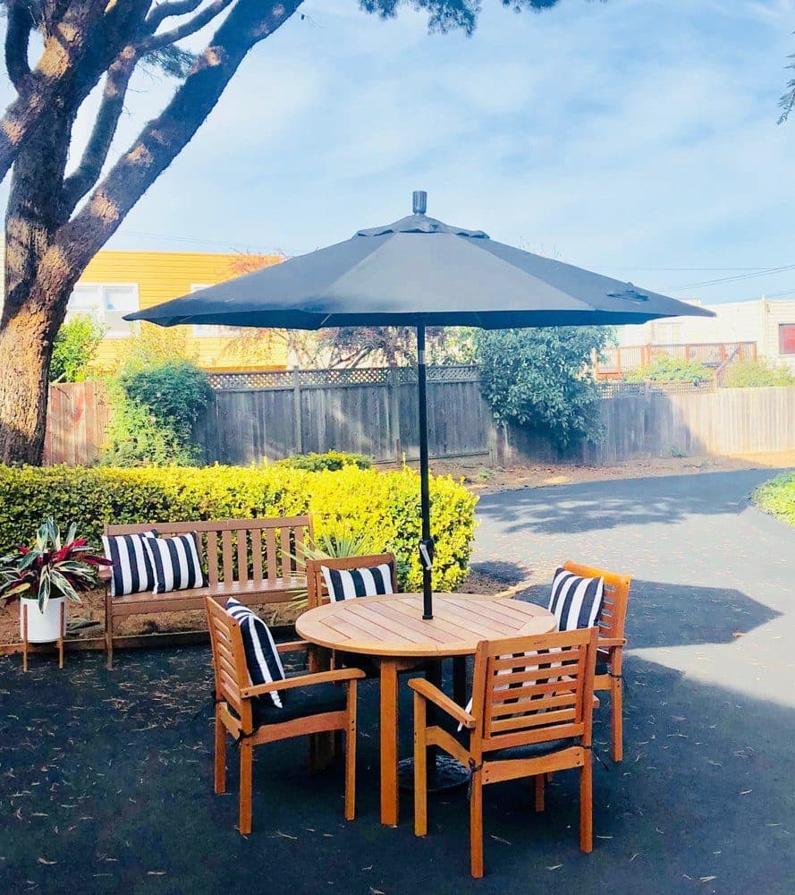 The back garden at Portola Gardens, a private planted patio with shaded seating under a large umbrella, enclosed by trees and fencing
